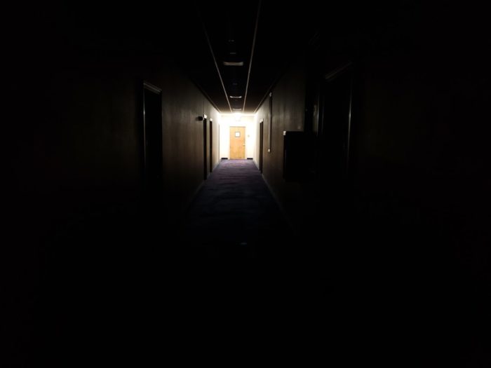 Dark hotel hallway with only one door lit at the far end.