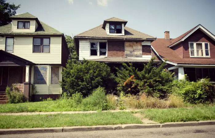 Three houses in a Detroit neighborhood. One of the houses is boarded up and has three broken windows. The front yard is overgrown and the bushes cover the view of the front porch.