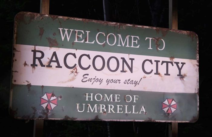 A worn-down sign that reads "Welcome to Raccoon City. Enjoy your stay! Home of Umbrella", adorned with two Umbrella logos.