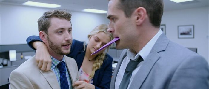 Two men and a woman talk in a close office space while one man holds an envelope in his mouth.