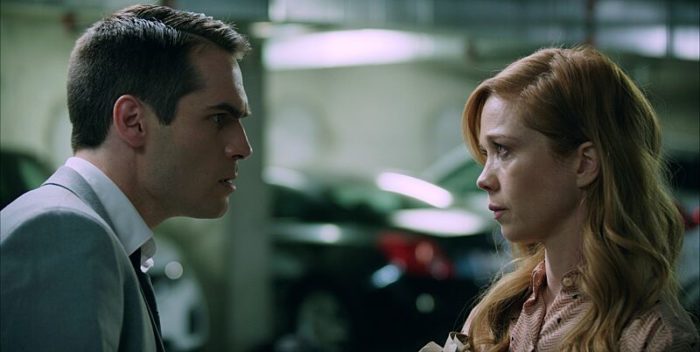 A man and a woman talk in a close frame in a parking garage.