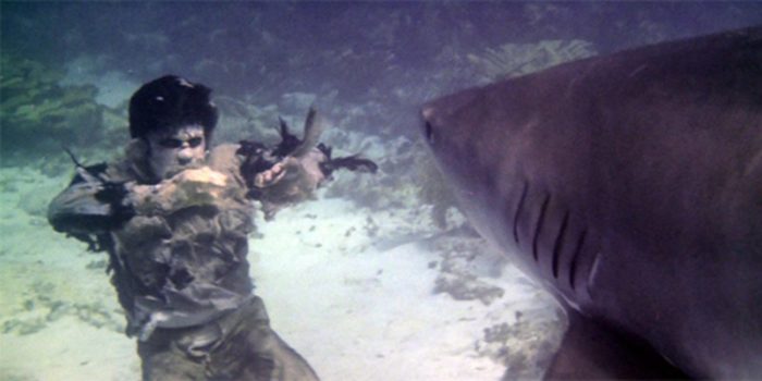 A zombie with rotted skin and tattered clothes floats underwater as a shark swims toward it.