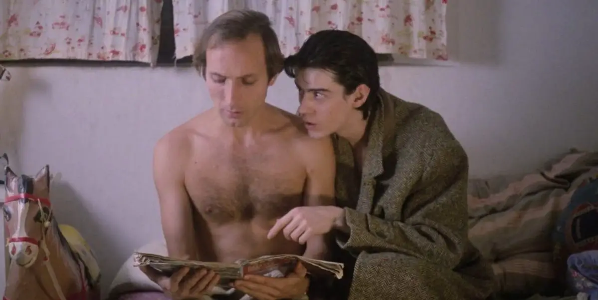 José and Pedro sit on a bed looking at a magazine and Pedro is pointing to something inside of it in Arrebato