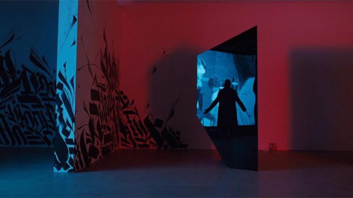 A red room with a projector screen in the background, the silhouette of a man with a hook in place of one hand standing in front of the projector