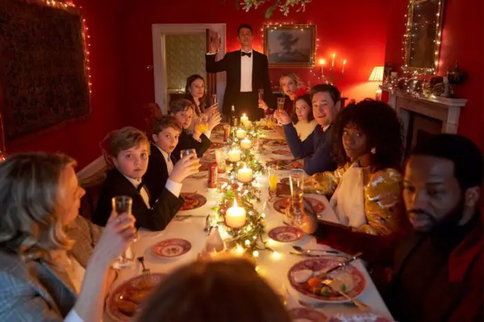 The cast of Silent Night raises a toast while sitting at a long Christmas themed dinner table. MAtthew Goode stands at the head raising his glass.