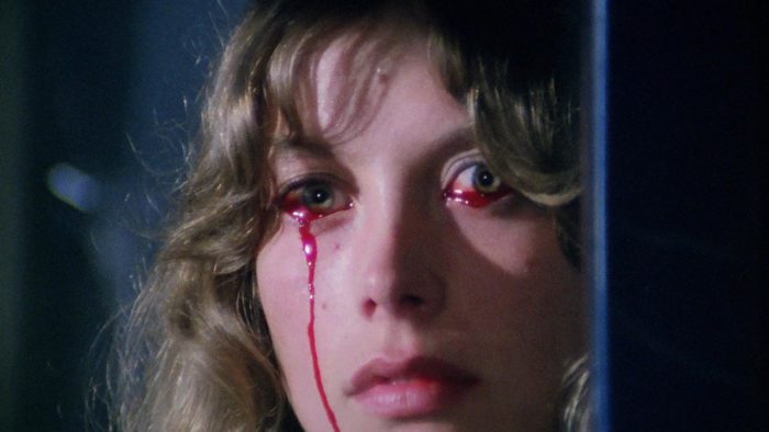 A close shot of a woman's face, her eyes open; a trail of blood drips from her right eye and down her cheek, while a drop of blood forms on the lower lid of her other eye.