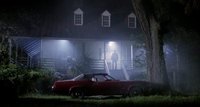 A long shot of a man seen from behind standing on a brightly lit porch at night, in the foreground at the bottom of the steps to the porch is a parked car; visible in the lights is a cloud of fog.