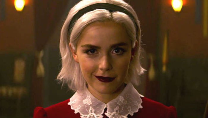 Close view of Sabrina with platinum hair, black headband, and dark lipstick wearing a half-smile.