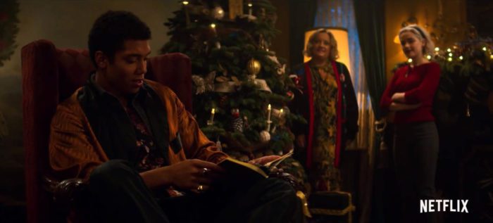 Ambrose reads a book in an armchair while Sabrina and Hilda look on near the Yule tree
