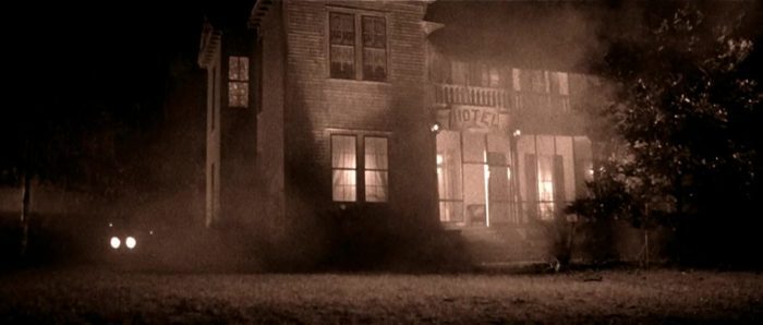 Seen at night, through fog and from a low angle, a two story house with a "Hotel" sign over the front porch; interior lights are visible through the first floor windows, while the headlights of a car can be seen approaching in the darkness to the left of the house.