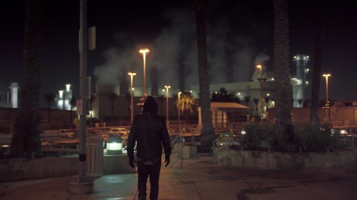 A hooded figure in the night stands on a sidewalk overlooking a parking lot