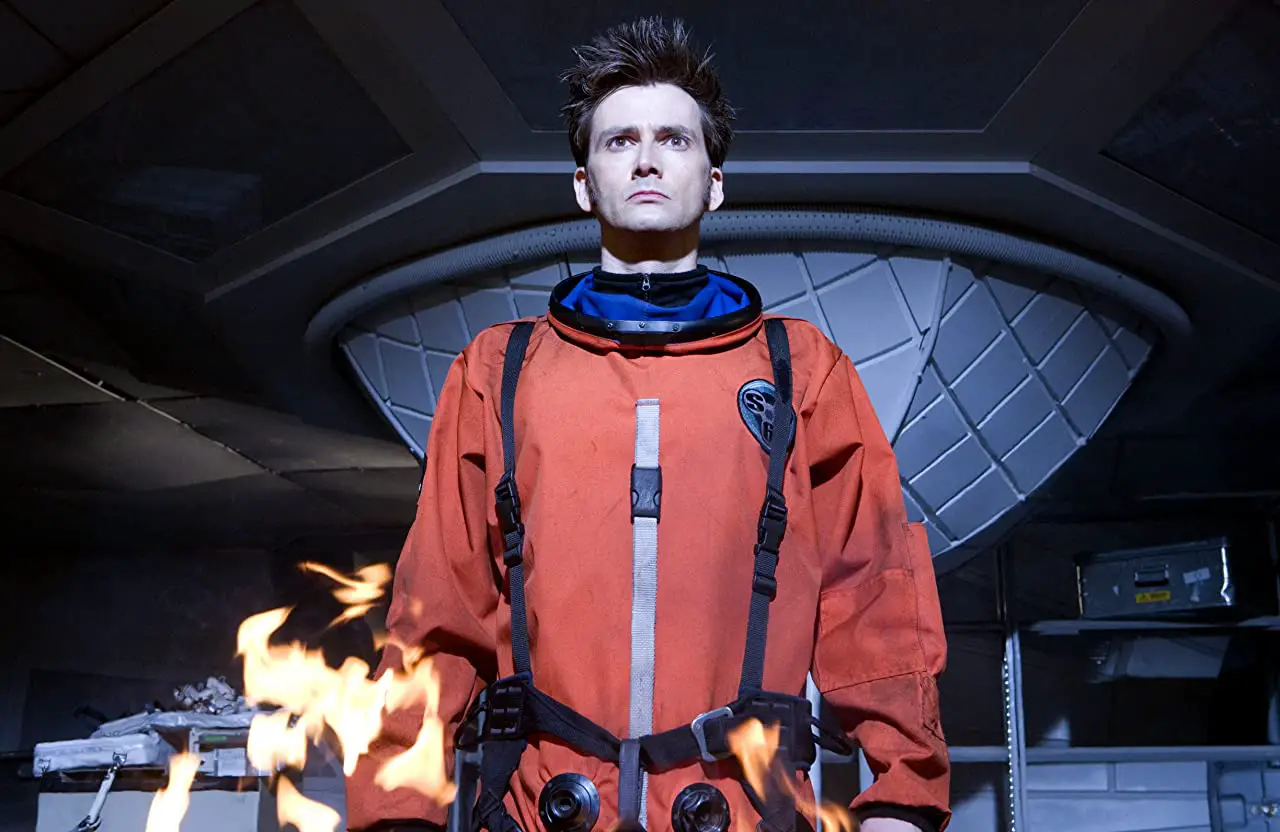 The Tenth Doctor, wearing an orange spacesuit and standing in front of a fire