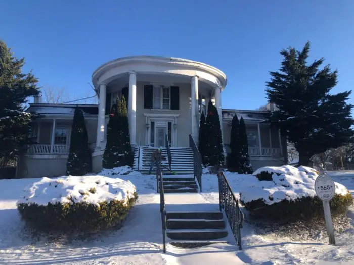 A two-story mansion with an octagon-shaped porch with tall columns sits dusted in snow.