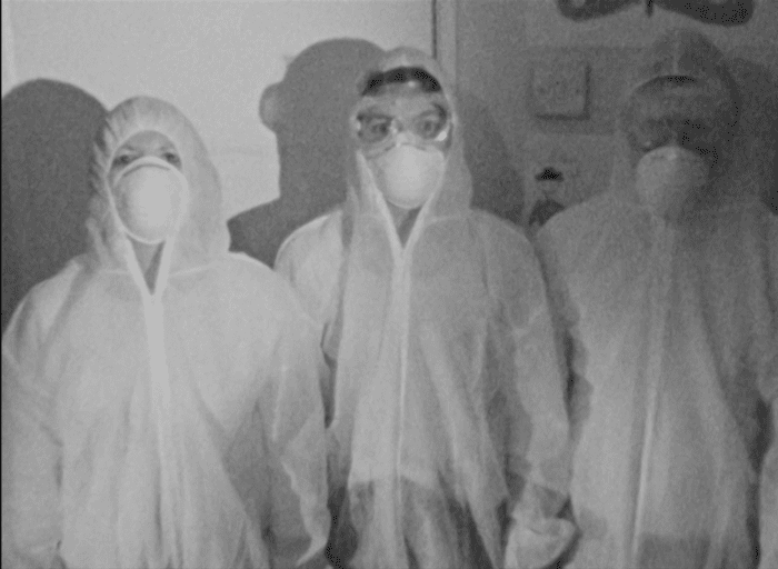 three people in medical garb stand facing the camera. the footage is in black and white.