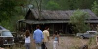 Four people, two men and two women, approach a rundown house in the woods.