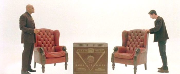 Morpheus and Neo talk between two identical leather seats and a tv in an otherwise white liminal space in The Matrix