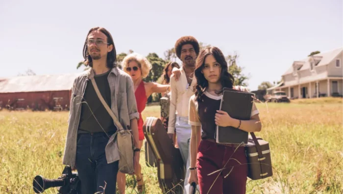 A group of actors carrying equipment walk through a field in X