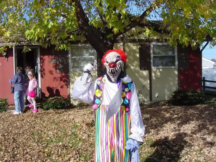 A monstrous-looking clown stands outside of a house holding a knife.