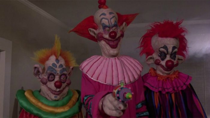 Three very cartoonish clowns stand looking at someone or something off-camera. The middle clown points a gun at unseen target.