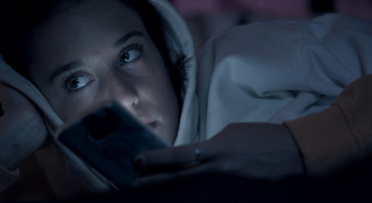 A screenshot from Ego shows Paloma (Maria Pedraza)laying in bed, idly holding her phone, lit by the light of the screen, and looking to the left