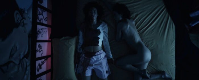 A screenshot from Ego shows Paloma (Maria Pedraza) laying on a bed in a bloodstained shirt and shorts with a knife, her nude double beside her