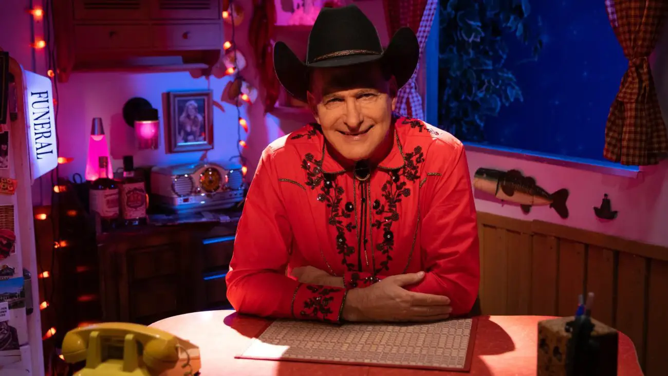 Promo shot of Joe Bob in the trailer set, leaning on a table, smiling