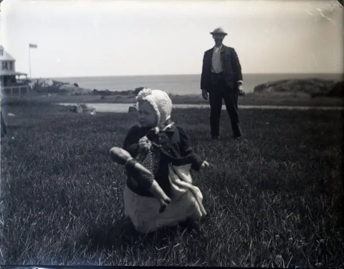 A child dressed in old fashioned 19th century clothing stands in a field as a man looks on in the background.