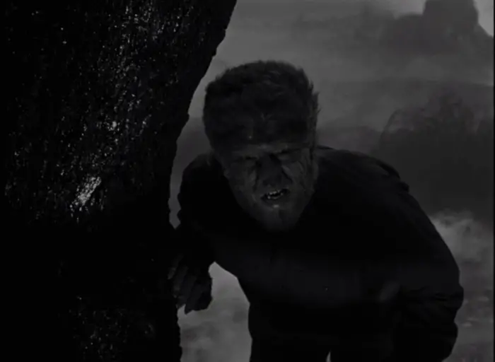 A wolfman leans himself against a tree with a clawed hand and looks suspiciously into the camera