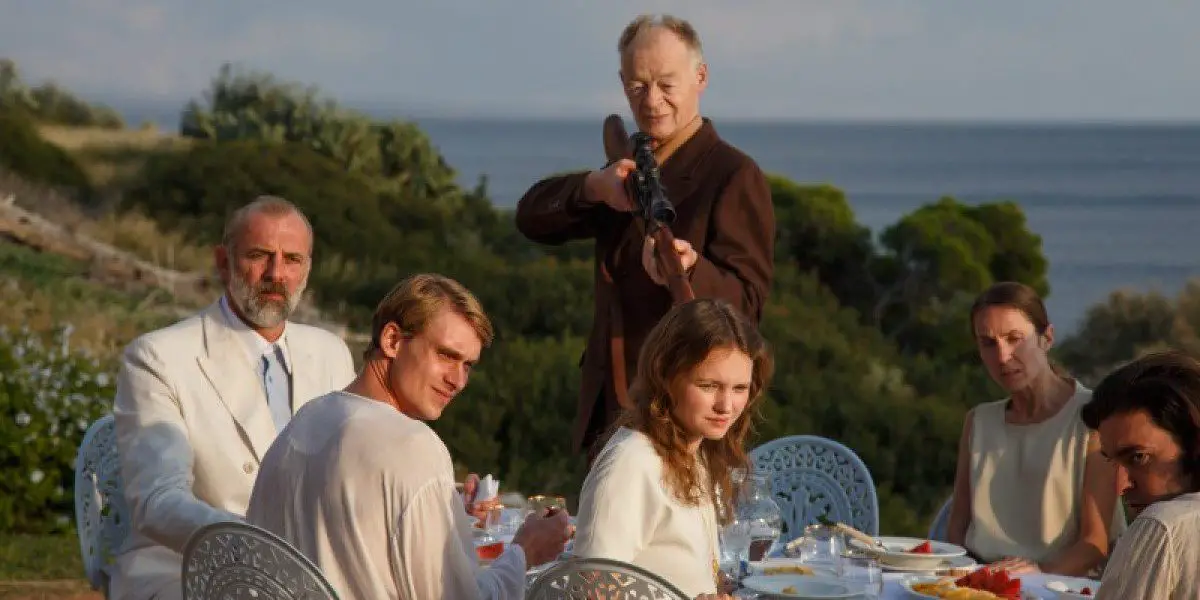 An oceanside banquet where all the guests are turned toward the camera, one of which is pointing a rifle in that direction.