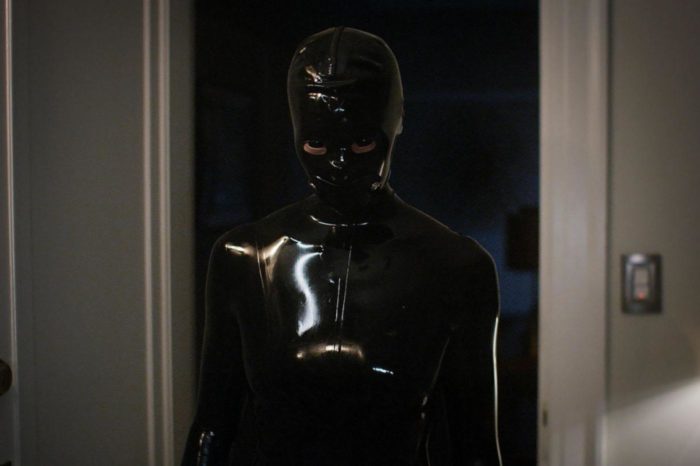 A mysterious figure stands fully covered in a black shiny latex suit, staring at the camera.