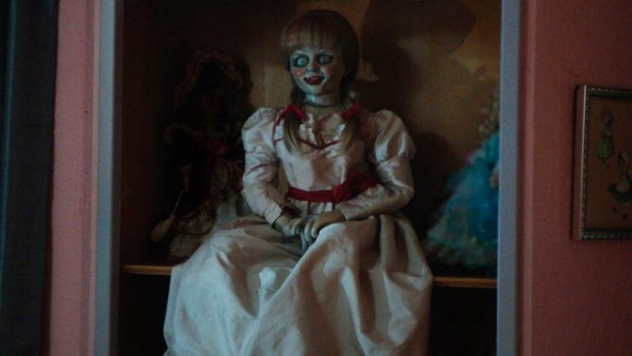 A vintage doll in a white dress sits on a shelf in a dark room.