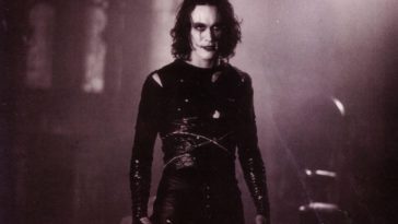 A man dressed in black wearing white face makeup and black makeup around his eyes stands in the dark.