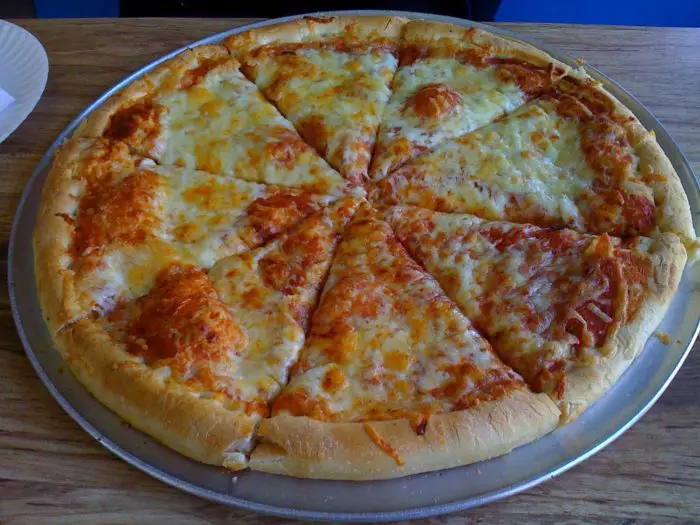A plain cheese pizza sits on a round metal tray on a wooden table.