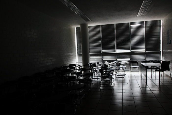 A mostly dark classroom with some harsh white light breaking through the blinds to show desks in silhouette.