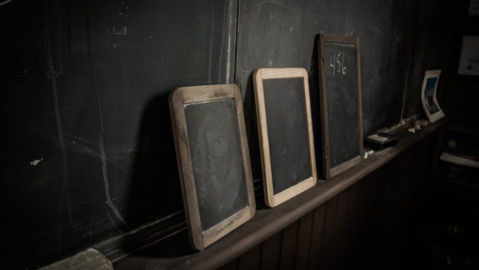 A dark photo of slates lined up against a chalkboard in an old fashioned classroom.