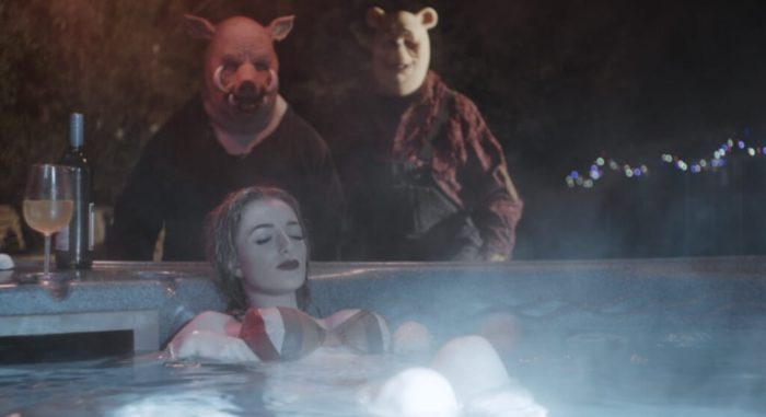 A woman relaxes in a hot tub as two people, dressed in animal costumes approach.