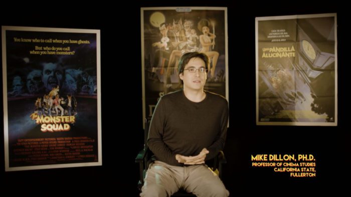 Mike Dillon, Ph.D., Professor of Cinema Studies for California State, Fullerton, speaks in the documentary, "Wolfman's Got Nards" (2018), with movie posters behind him, including "The Monster Squad" (1987).
