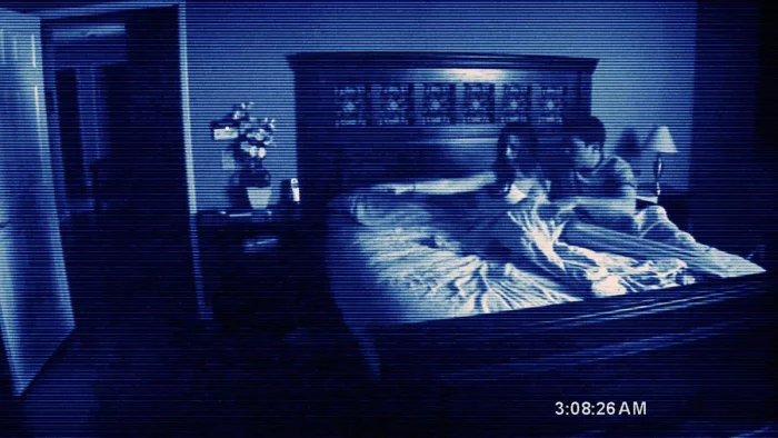 A static camera set up in a bedroom looks at a woman and man awake in bed after hearing a sound in the night