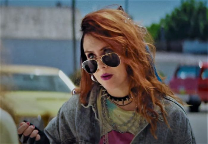 Caito Aase as Angie Pitarelli in the horror film Revealer, looking very punk wearing a denim jacket, sunglasses, and spiked collar.