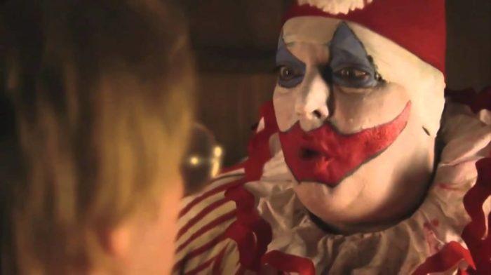 A closeup of a man in a clown costume and makeup talking to a man in the foreground facing away from the camera.