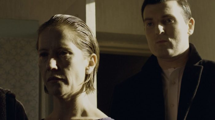 A woman with no eyes and a mystery man stand facing the camera next to an opened door