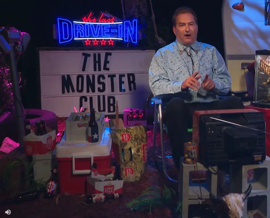 Joe Bob on The Last Drive-In set with a sign for The Monster Club behind him.