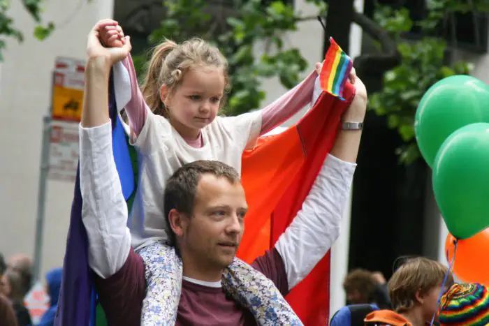 A father carries his daughter on his shoulders, an LGBTQ+ pride flag is draped around her shoulders and she holds a mini-pride flag in her hand.
