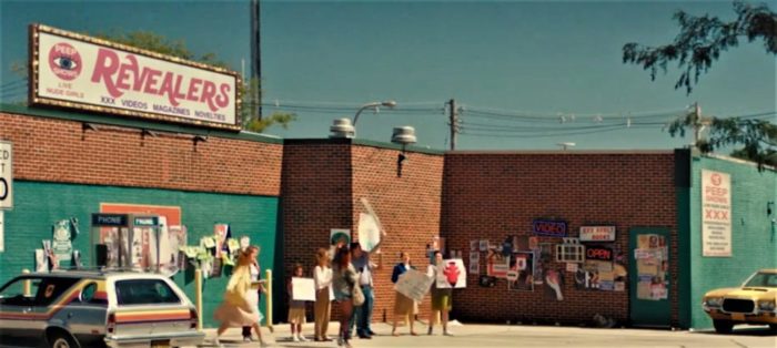 A billboard over a scummy building states Revealers, the strip club in the horror film Revealer. Out front are several religious protesters.