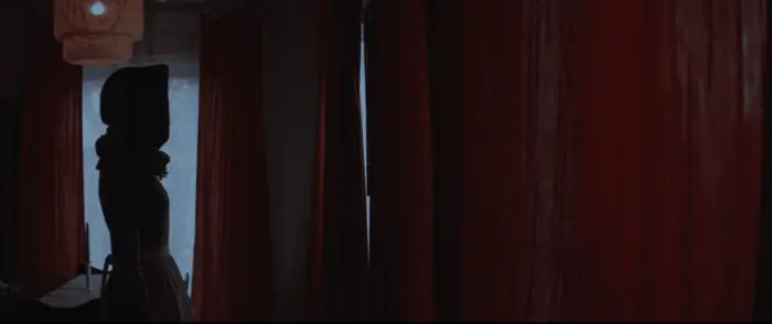 A hooded figure stands against a backdrop of red curtains in The Unsettling