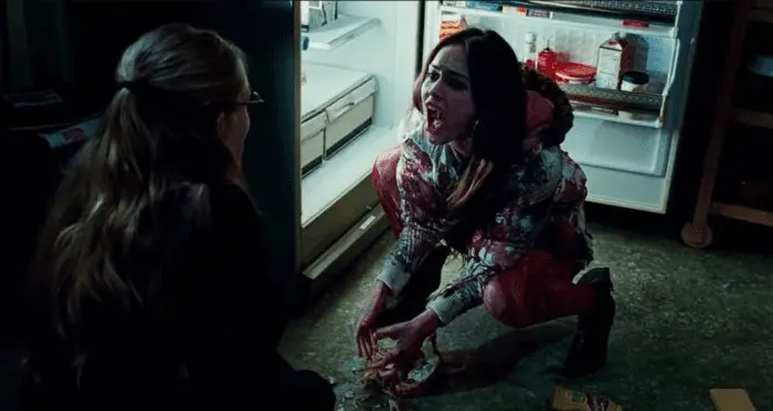 A dark shot of two young women crouched on the floor in front of an open refrigerator. One is covered in blood.