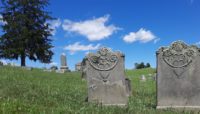 Two tombstones from the 18th century sit quietly in a grave yard against a blue sky.