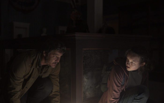 Pedro PAscal and Bella Ramsey duck in the light of a dark location in The Last of Us tv series.