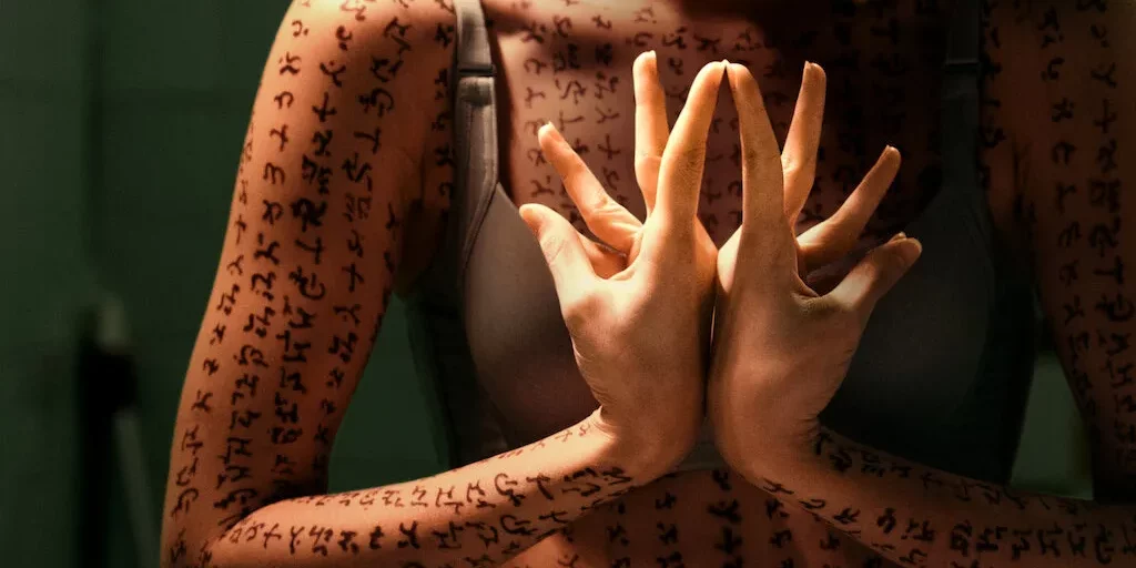A woman covered in runes, her hands creating a flower shape in prayer gesture