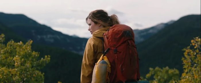 Joy stands in a yellow jacket and red backpack amid a beautiful mountainous backdrop